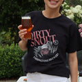A woman standing outdoors in a garden setting, wearing a black graphic t-shirt with pink and white text that reads 'DIRTY SNOUT' and 'FARRINGTONS BREWERY', paired with a white wrap skirt, while holding a pint of beer.