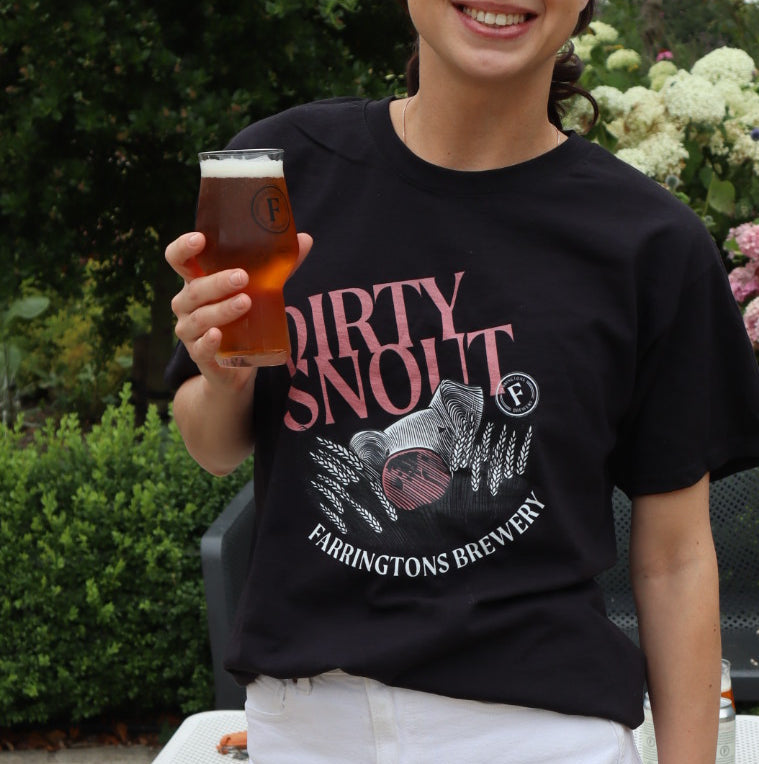 A woman standing outdoors in a garden setting, wearing a black graphic t-shirt with pink and white text that reads 'DIRTY SNOUT' and 'FARRINGTONS BREWERY', paired with a white wrap skirt, while holding a pint of beer.